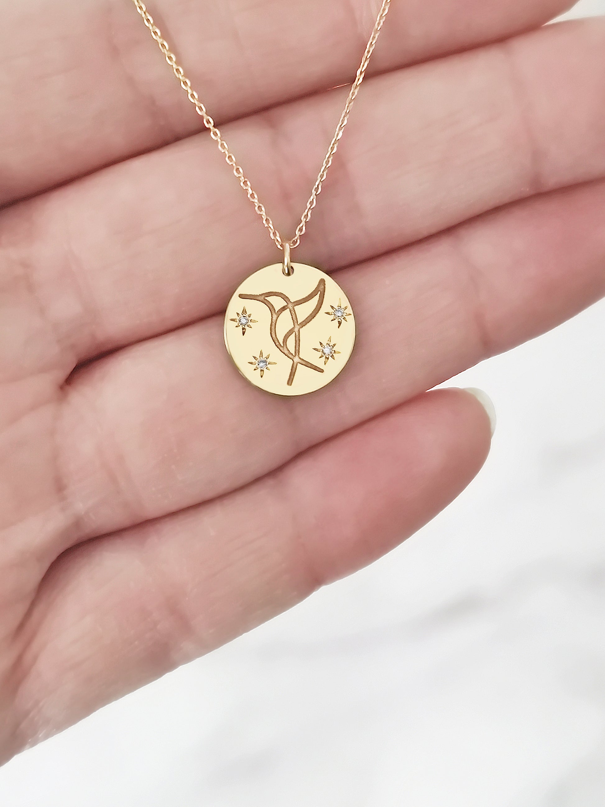 female hand holding gold necklace with 14mm disc pendant engraved with a hummingbird and set with four white diamonds with a star setting