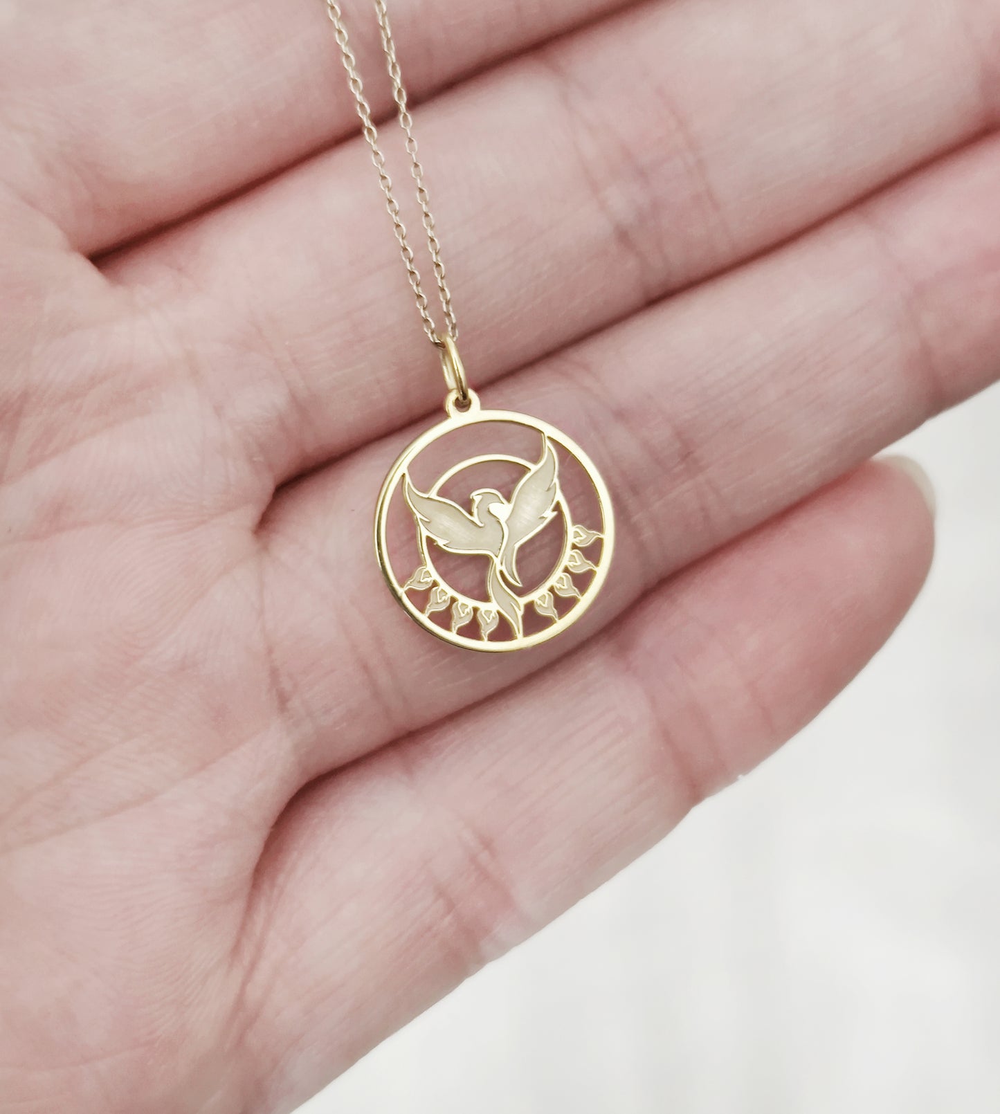 close up of a palm holding a yellow gold necklace with a cutout phoenix firebird pendant rising from flames for size reference