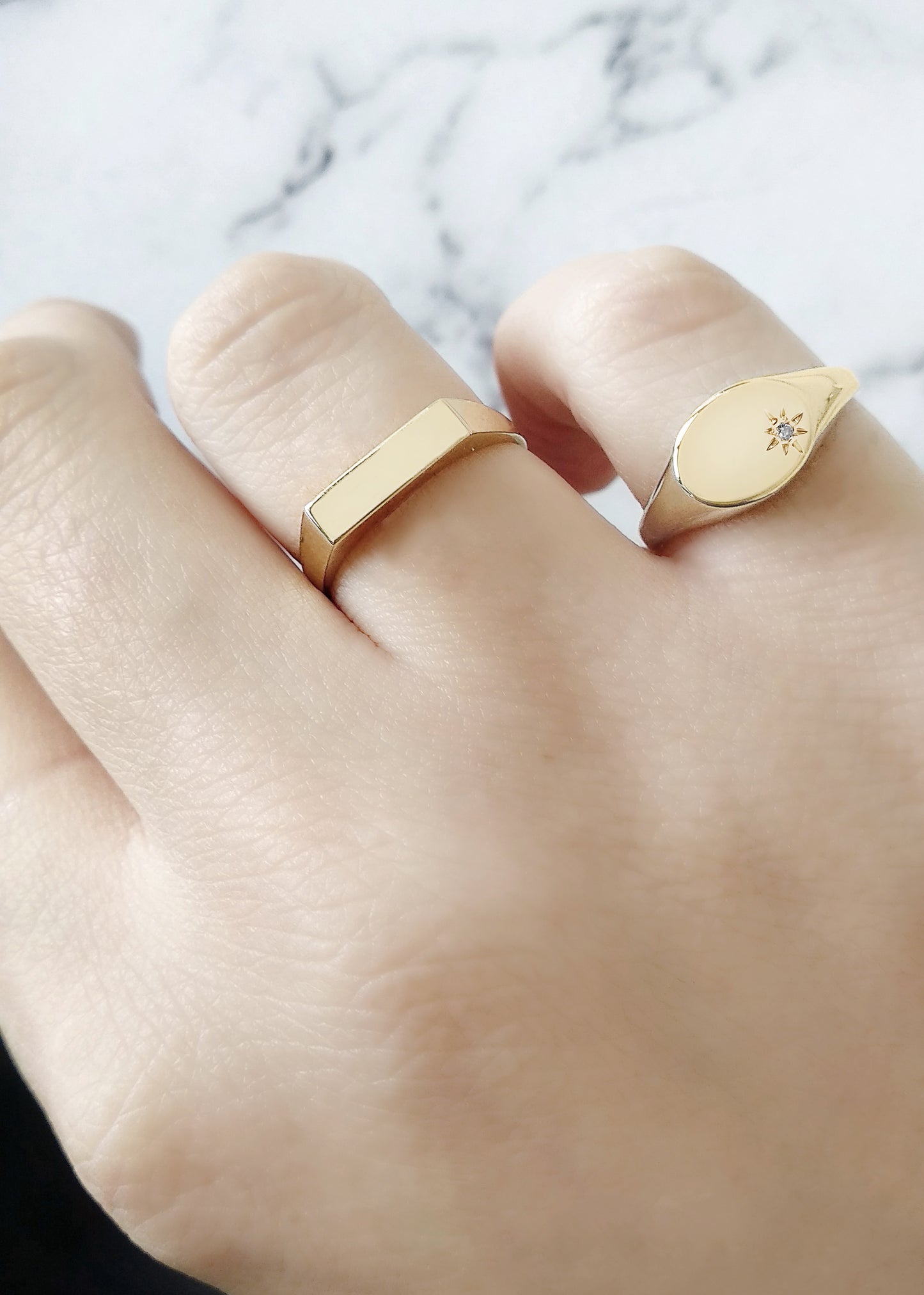 female hand wearing yellow gold oval signet ring set with a tiny diamond with a polaris star setting on her pinky and a thick bar signet ring on her ring finger