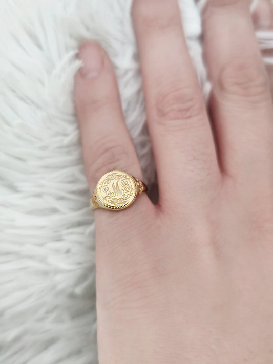 female hand wearing on her pinky a gold round signet ring with floral designs and engraved with three intertwined initials