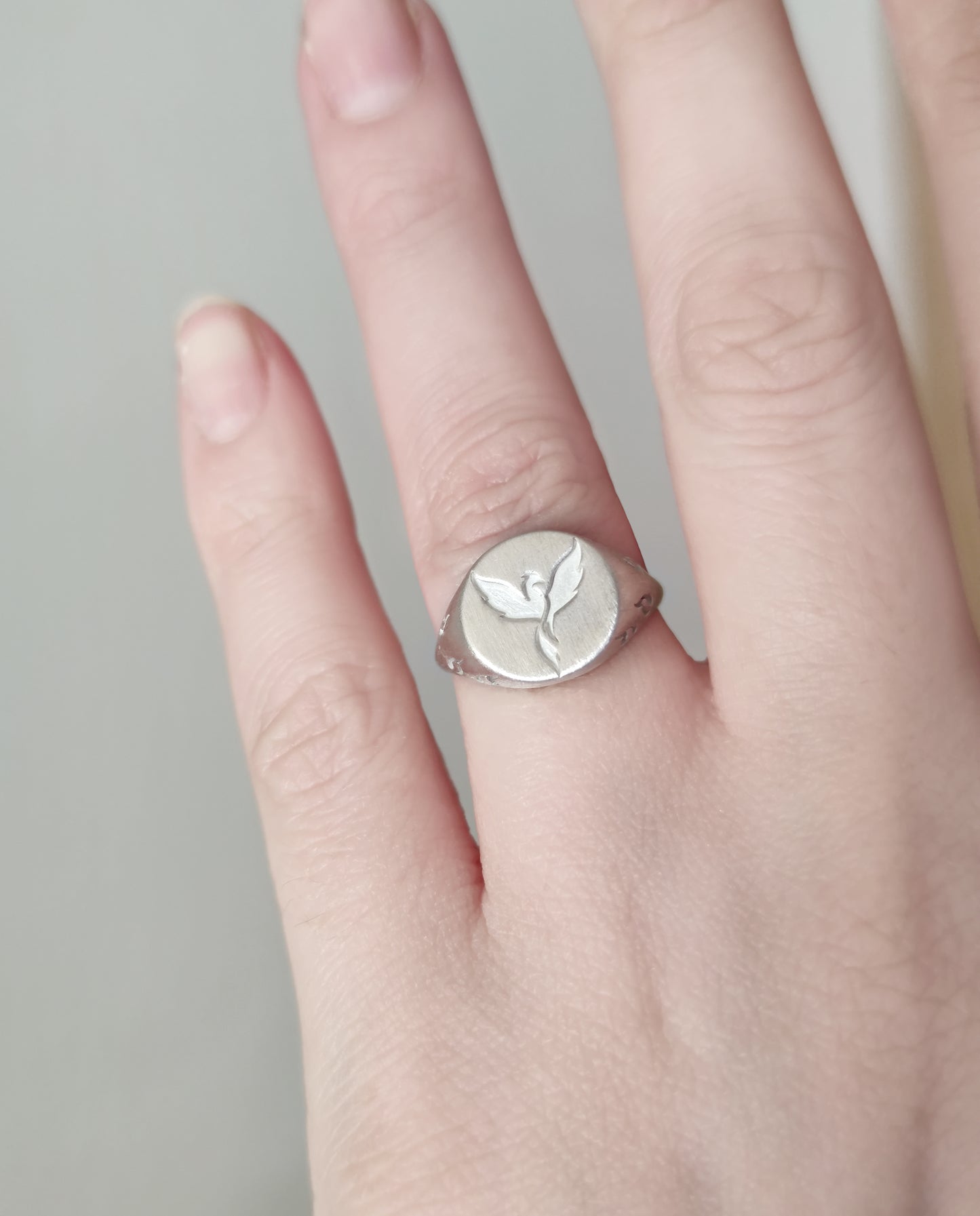 female hand wearing on the ring finger a white gold matte round signet ring engraved with a phoenix firebird rising with flame cutouts around the base of the ring's head