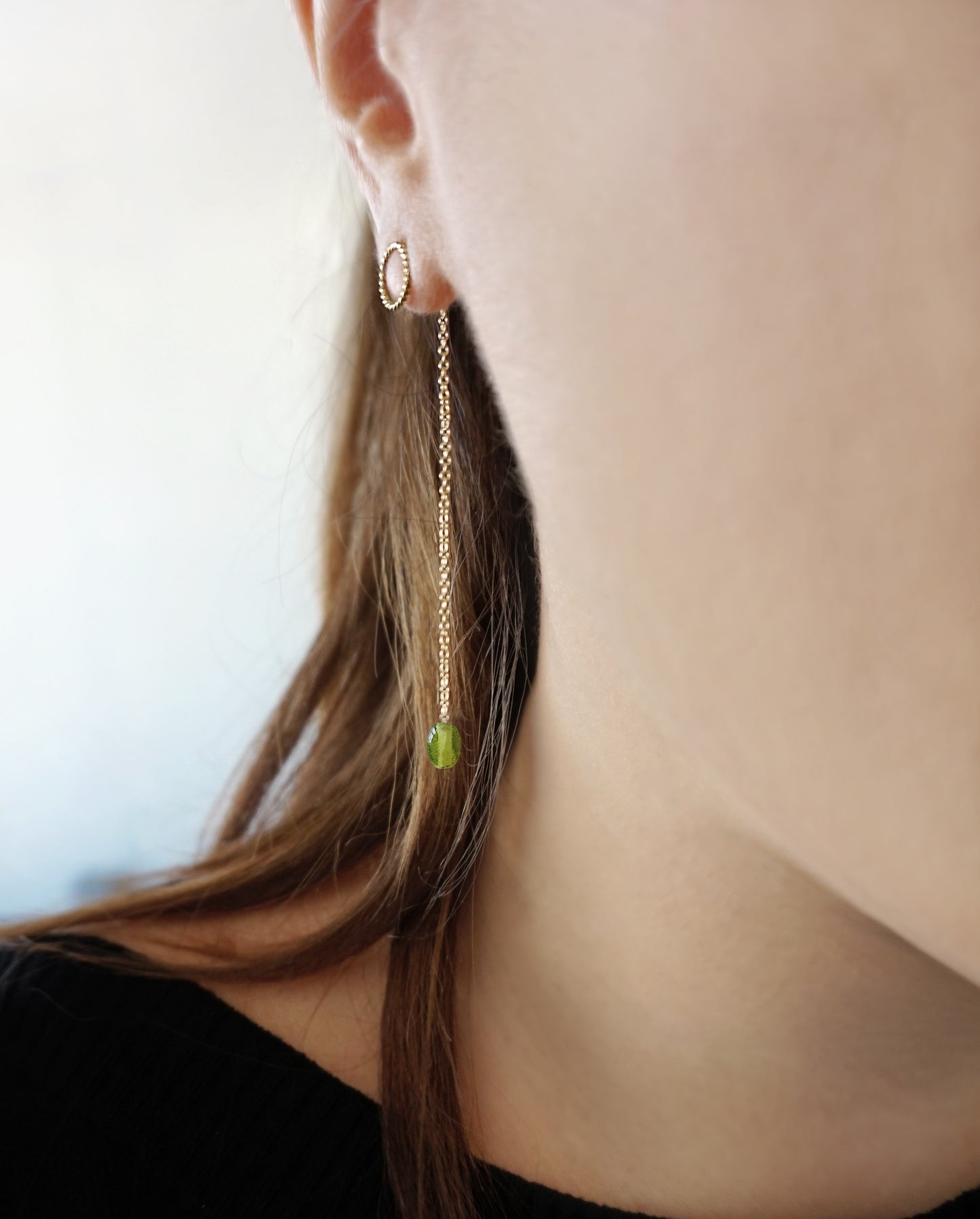 close up of a woman wearing yellow gold jacket earrings with a small twisted circle stud on the front of the earlobe and a long chain with a peridot bead at the end dangling on the back of the earlobe
