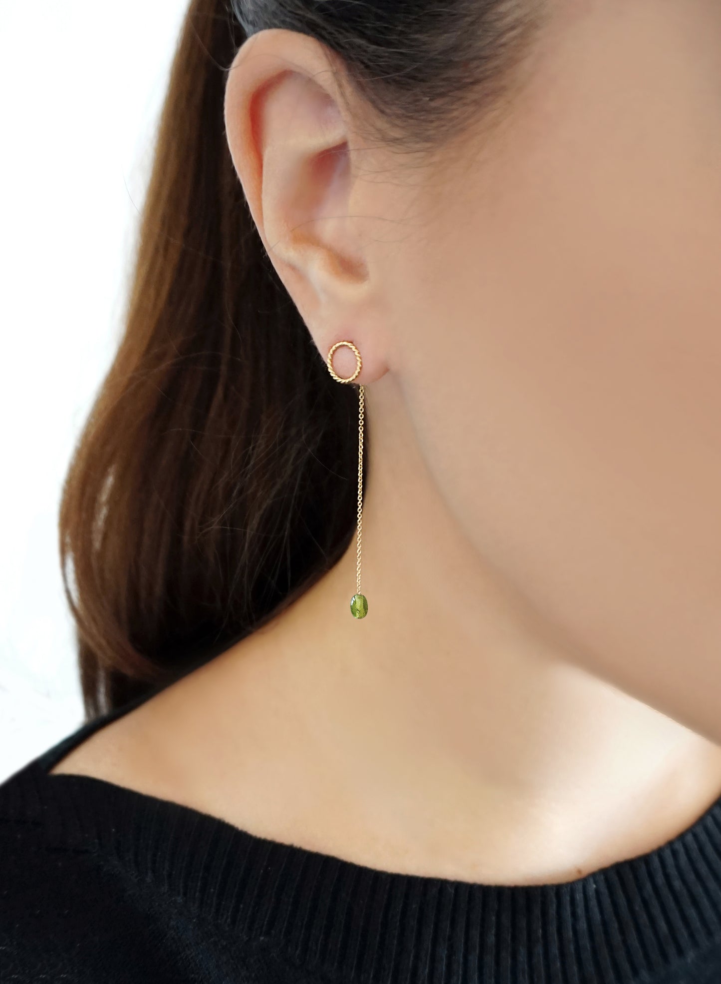 woman wearing yellow gold jacket earrings with a small twisted circle stud on the front of the earlobe and a long chain with a peridot bead at the end dangling on the back of the earlobe