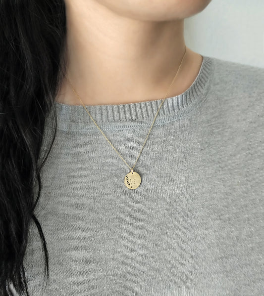 Gold necklace with a round hammered pendant worn by a female in a gray sweater.