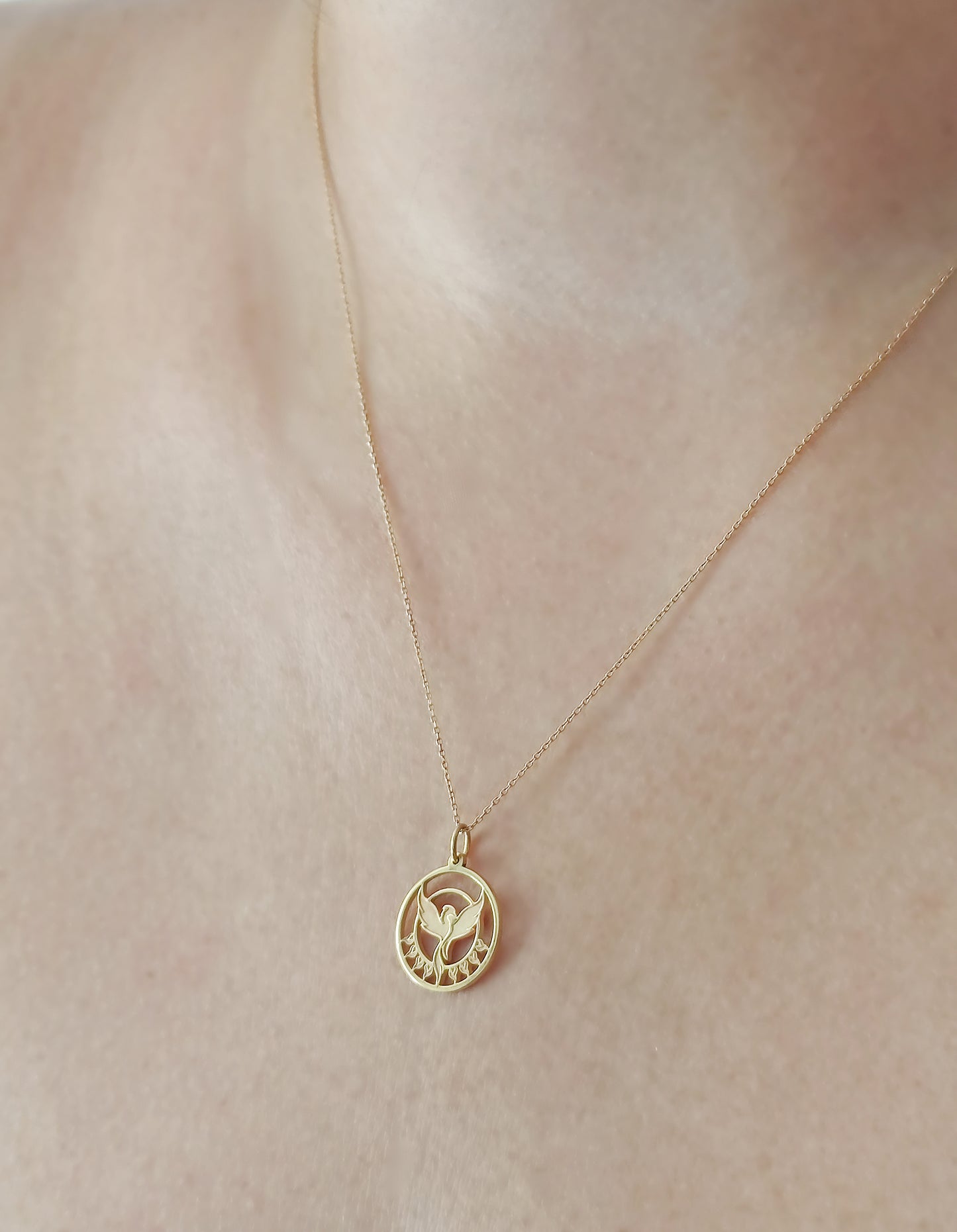 a woman's neck close up wearing a yellow gold necklace with a cutout phoenix firebird pendant rising from flames