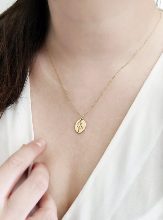 woman wearing a solid yellow gold oval pendant necklace engraved with a Snowdrop the birth flower of January