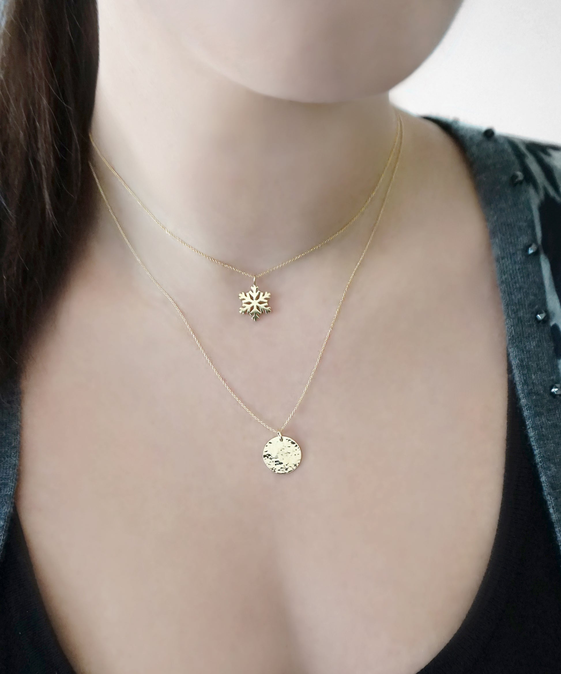 woman wearing gold necklace with a small pendant in the shape of a snowflake layered with a longer necklace with a hammered disc