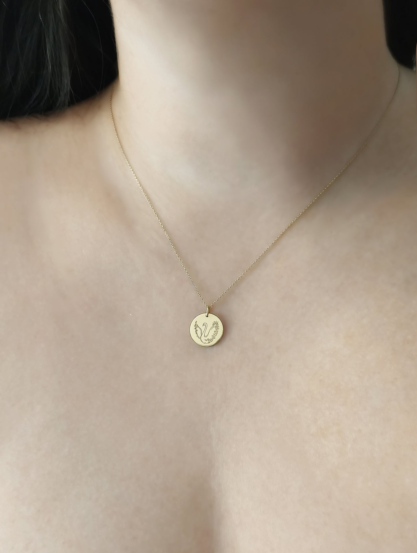close up of a woman's neck wearing a gold necklace with a 14mm disc pendant engraved with a swan