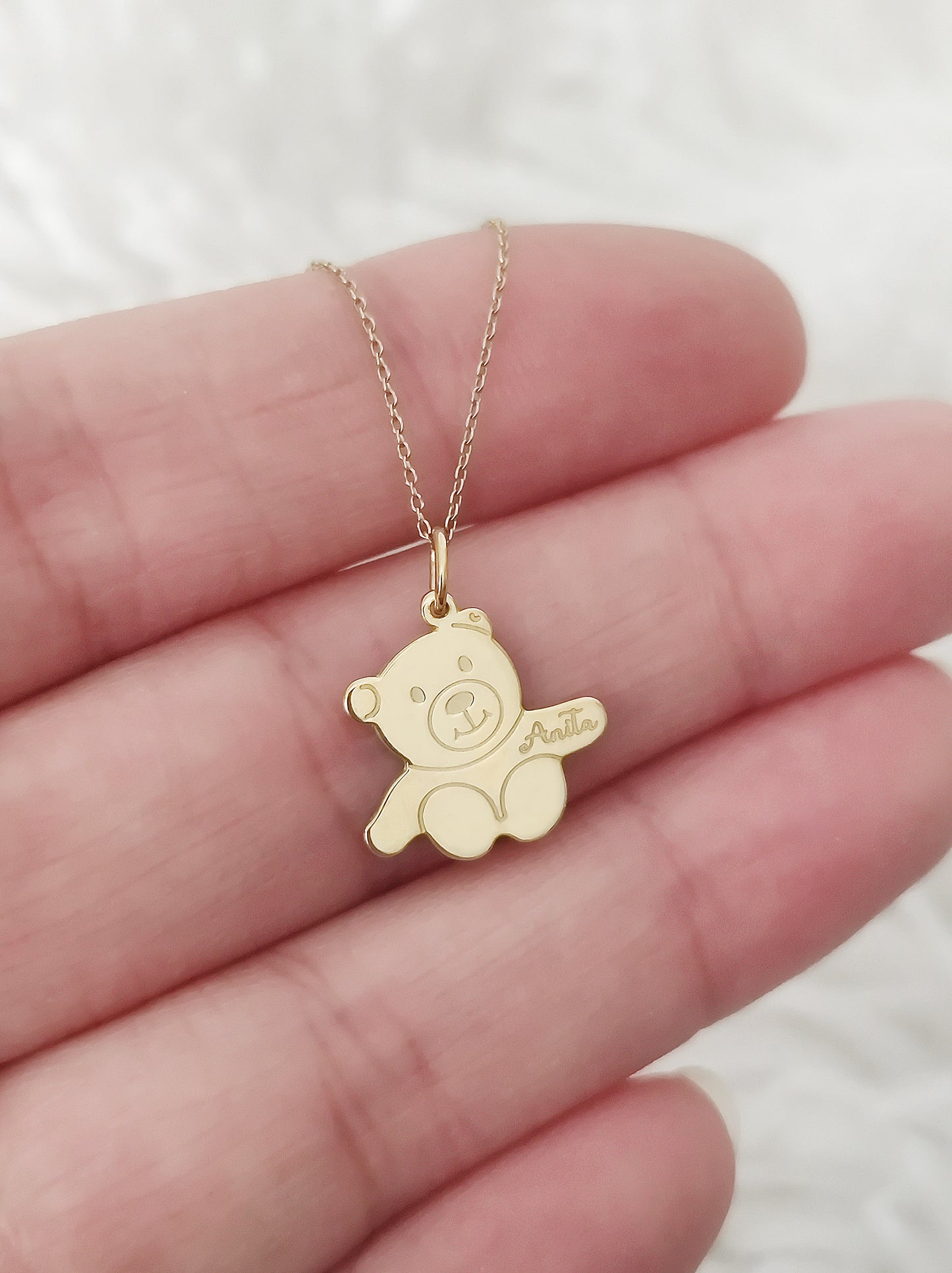 close up of a palm holding a gold necklace with a small teddy bear shaped pendant with open arms engraved with a baby's name for size reference