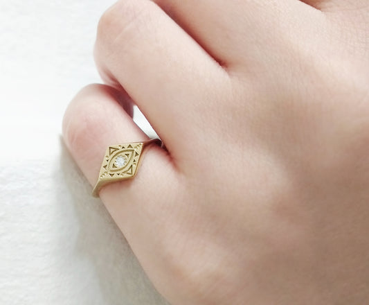 woman's pinky finger wearing yellow gold rhombus signet ring engraved with a geometric evil eye and set with a tiny white diamond in the center