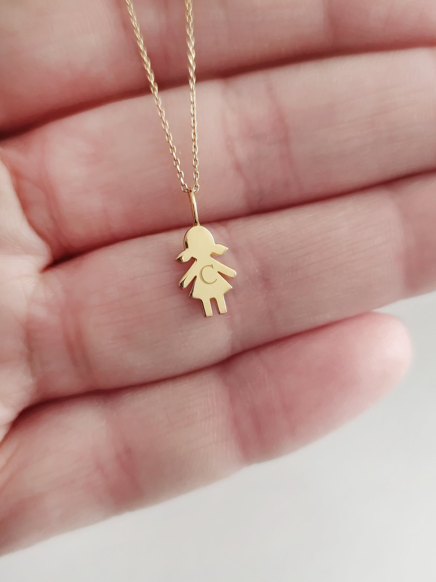 close up of a palm holding a gold necklace with a tiny girl outline charm engraved with an initial for size reference