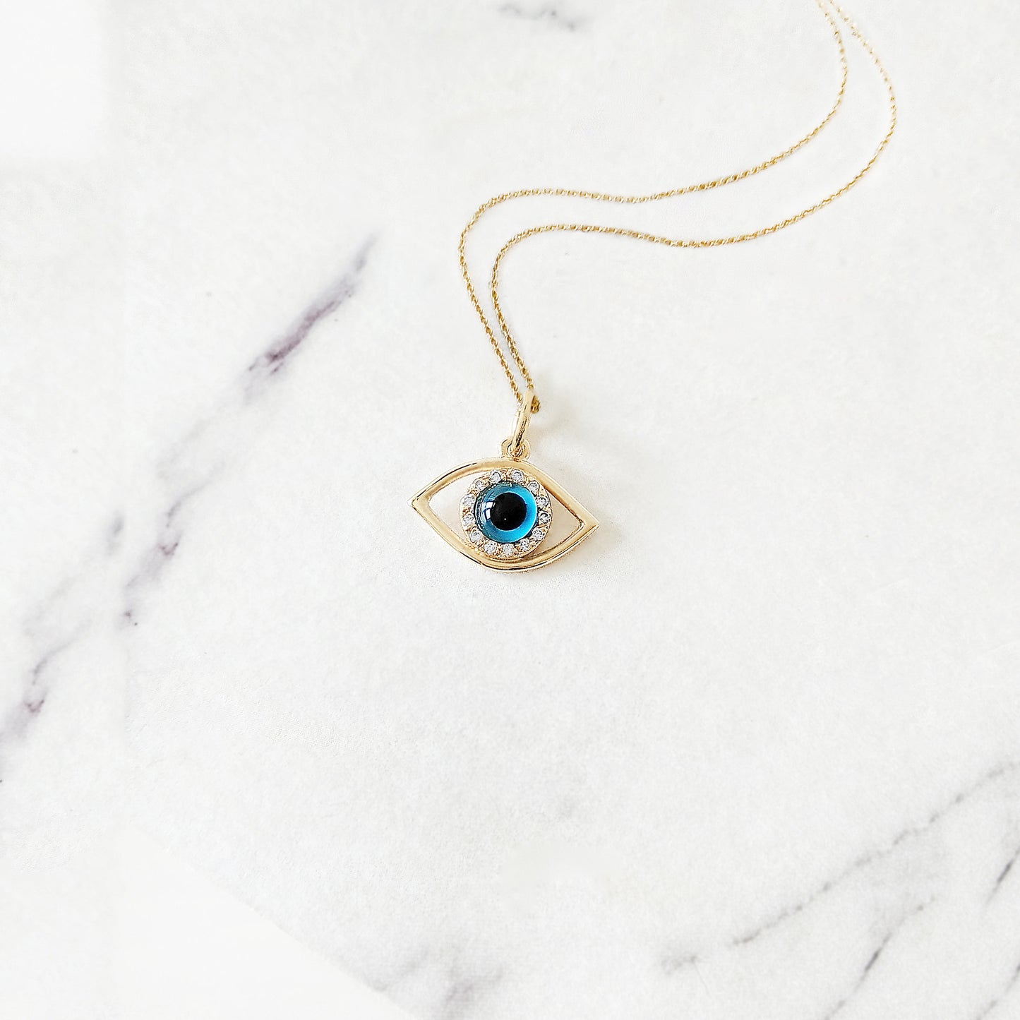 Yellow gold necklace with an eye-shaped pendant featuring a blue eye in the center surrounded by white diamonds on a marble surface