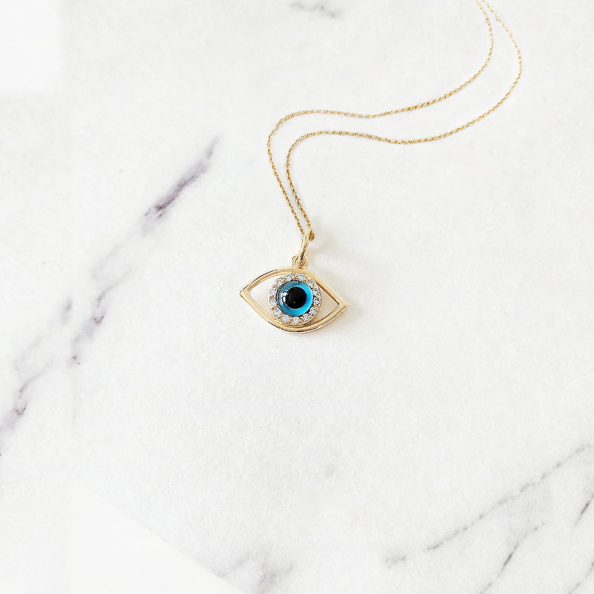 Yellow gold necklace with an eye-shaped pendant featuring a blue eye in the center surrounded by white diamonds on a marble surface