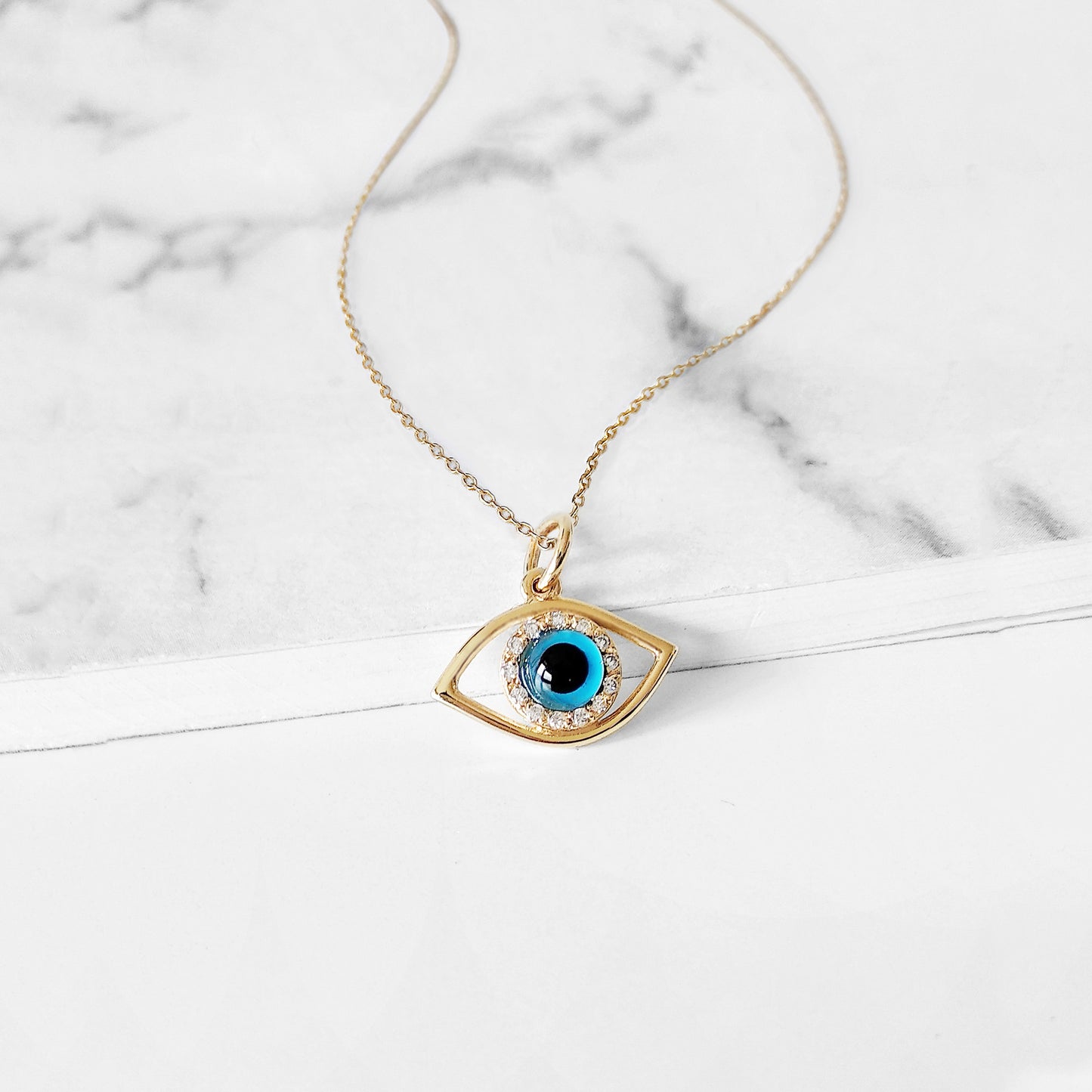 Gold necklace with an eye-shaped pendant featuring a blue eye in the center surrounded by white diamonds on a marble surface