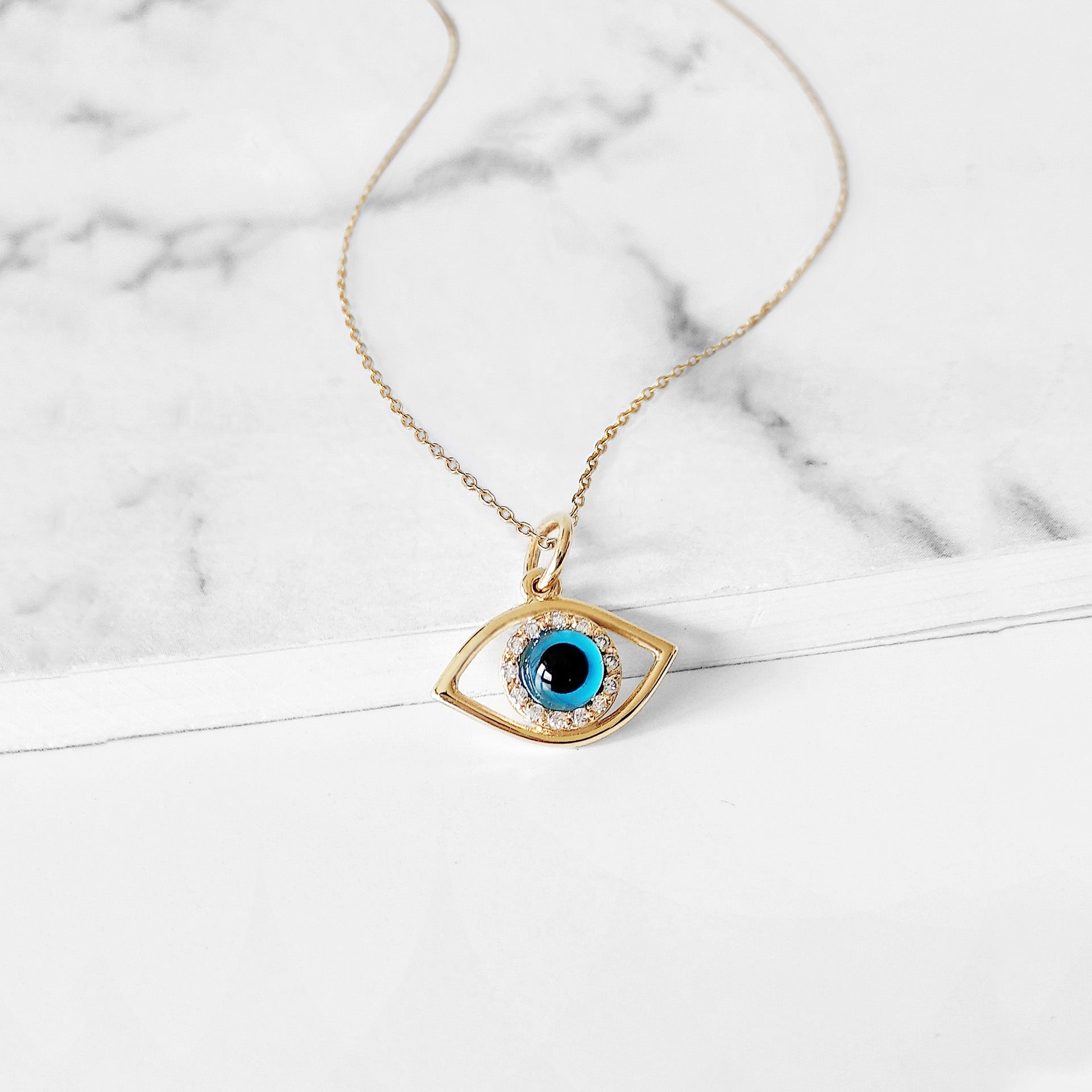 Gold necklace with an eye-shaped pendant featuring a blue eye in the center surrounded by white diamonds on a marble surface