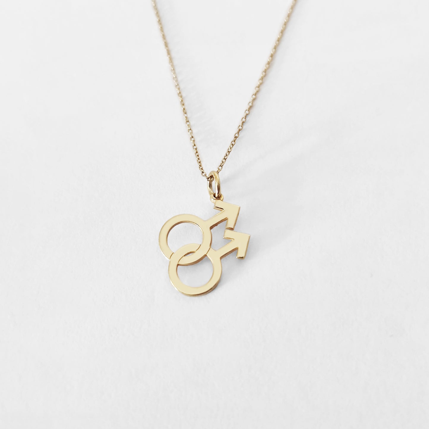 Gold necklace with a small double mars gay symbol pendant on a light gray background