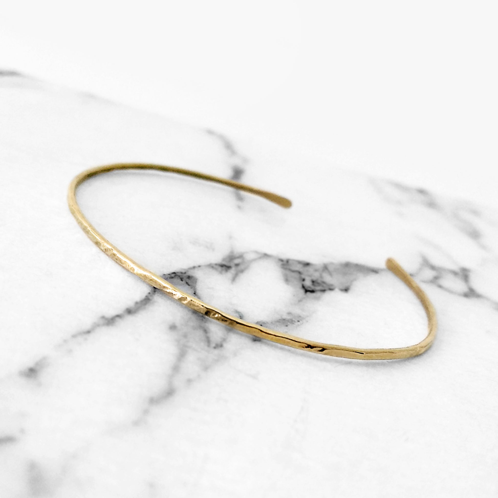 Back view of a thin gold hammered open bangle bracelet on a marble surface