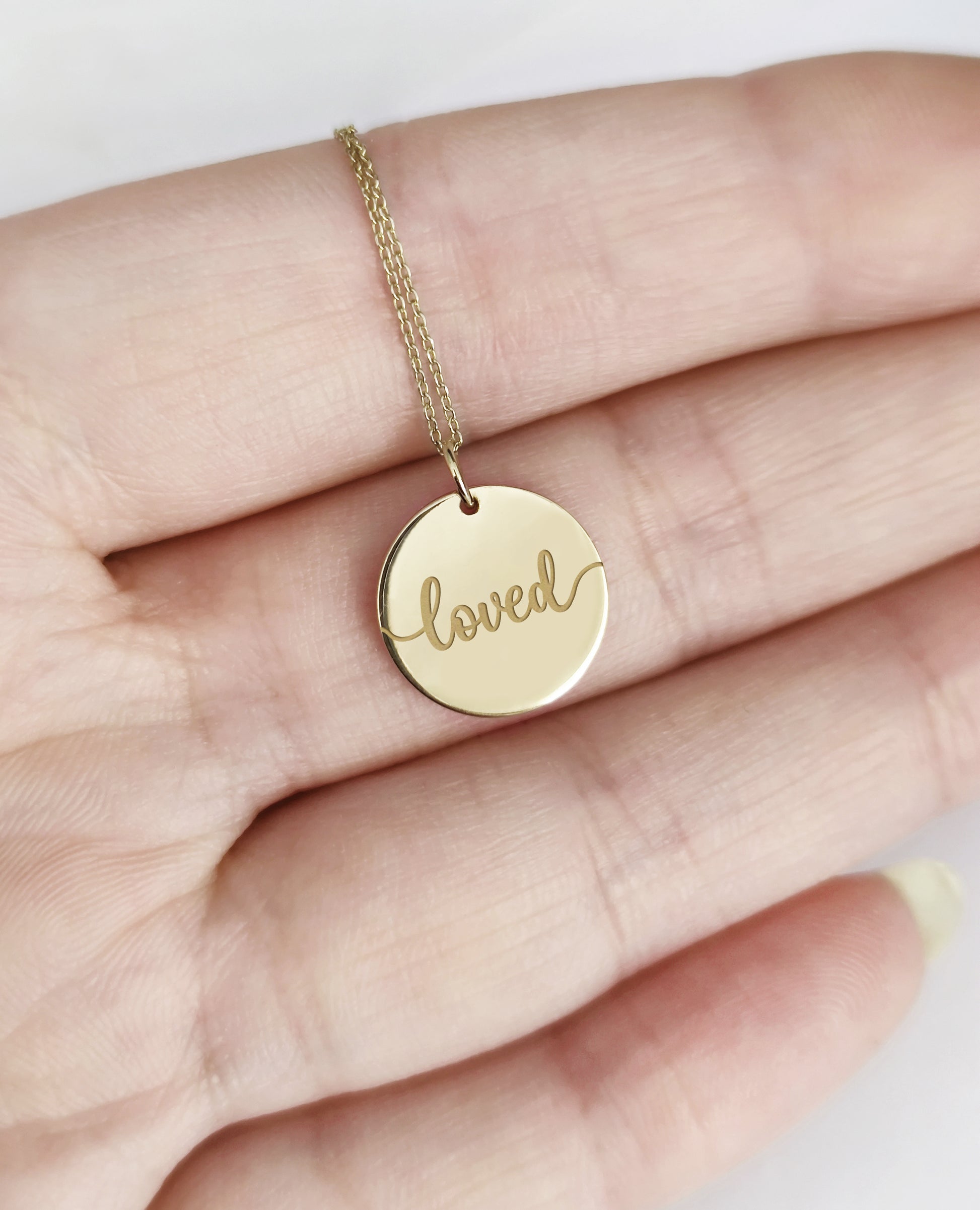 Gold necklace with a round pendant engraved with the word 'loved' held in a hand.