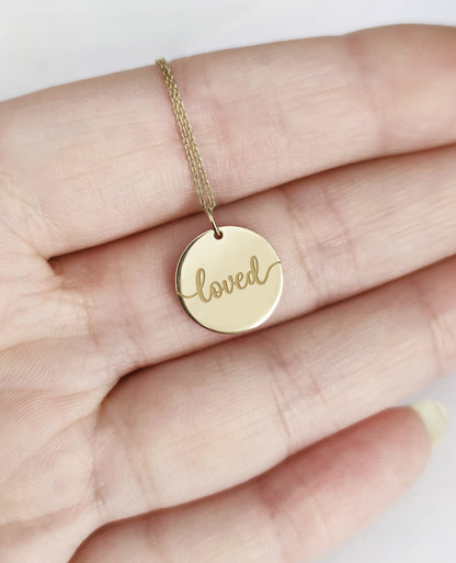 Gold necklace with a round pendant engraved with the word 'loved' held in a hand.