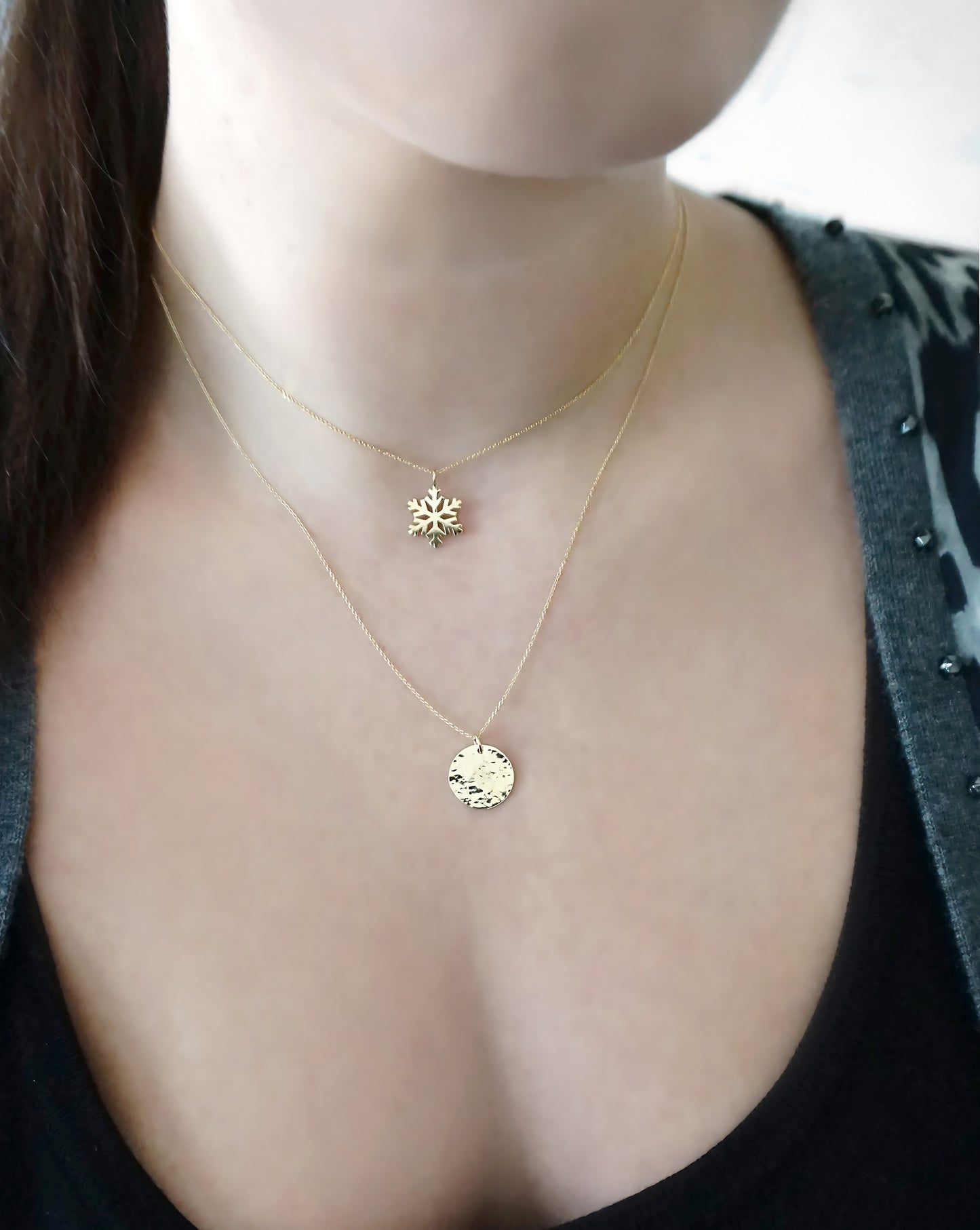 Close-up of a female wearing two gold necklaces one with a round hammered disc pendant and the other with a snowflake shaped pendant.