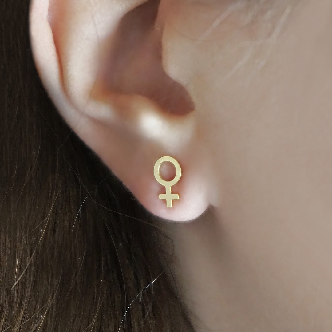 Gold female symbol earring worn on an ear with a neutral background