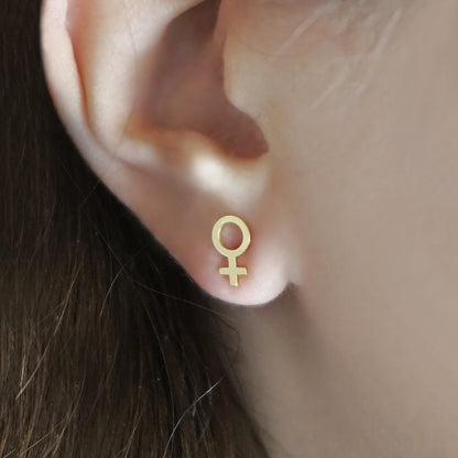 Gold female symbol earring worn on an ear with a neutral background