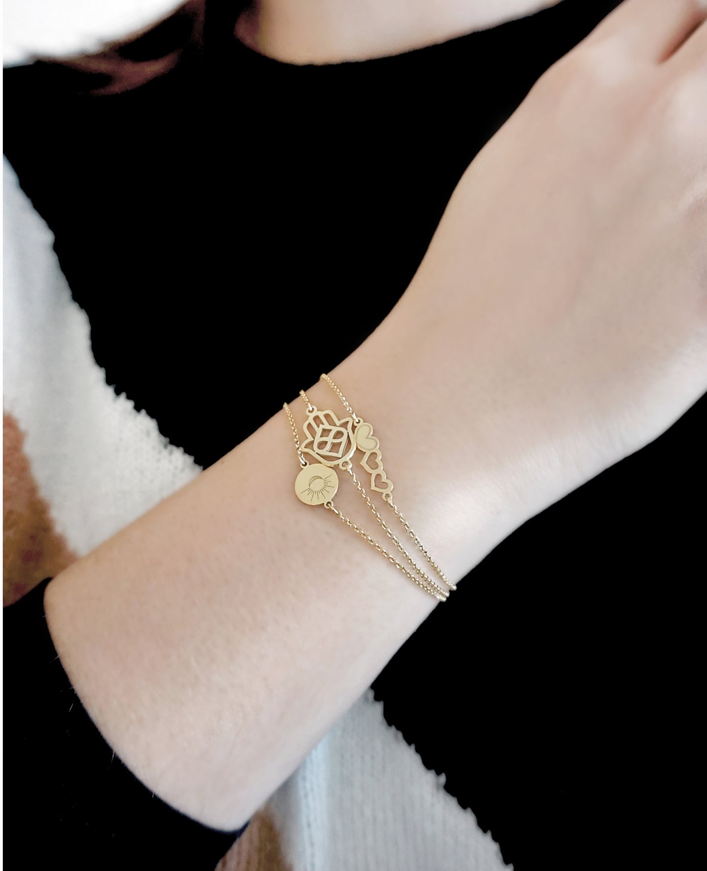 female wrist wearing three gold bracelets a sun and moon bracelet, a hamsa bracelet and a triple hearts bracelet
