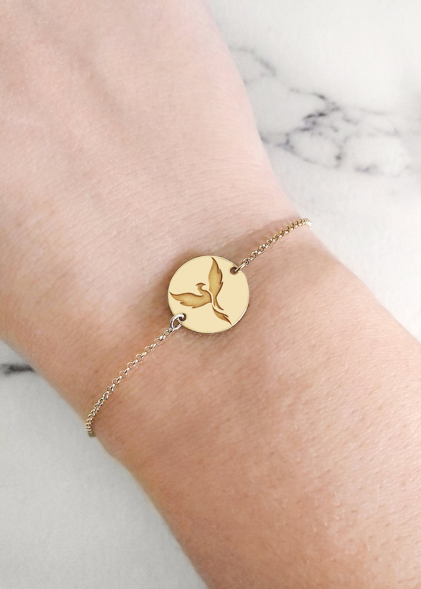 a woman's wrist wearing a  gold bracelet with a 14mm disc charm engraved with a phoenix firebird rising