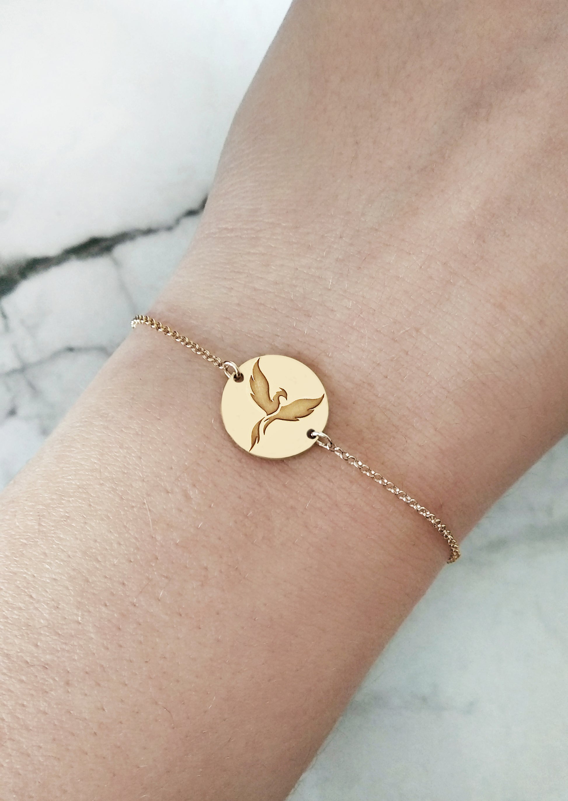 wrist of a woman wearing a gold bracelet with a 14mm disc charm engraved with a phoenix firebird rising