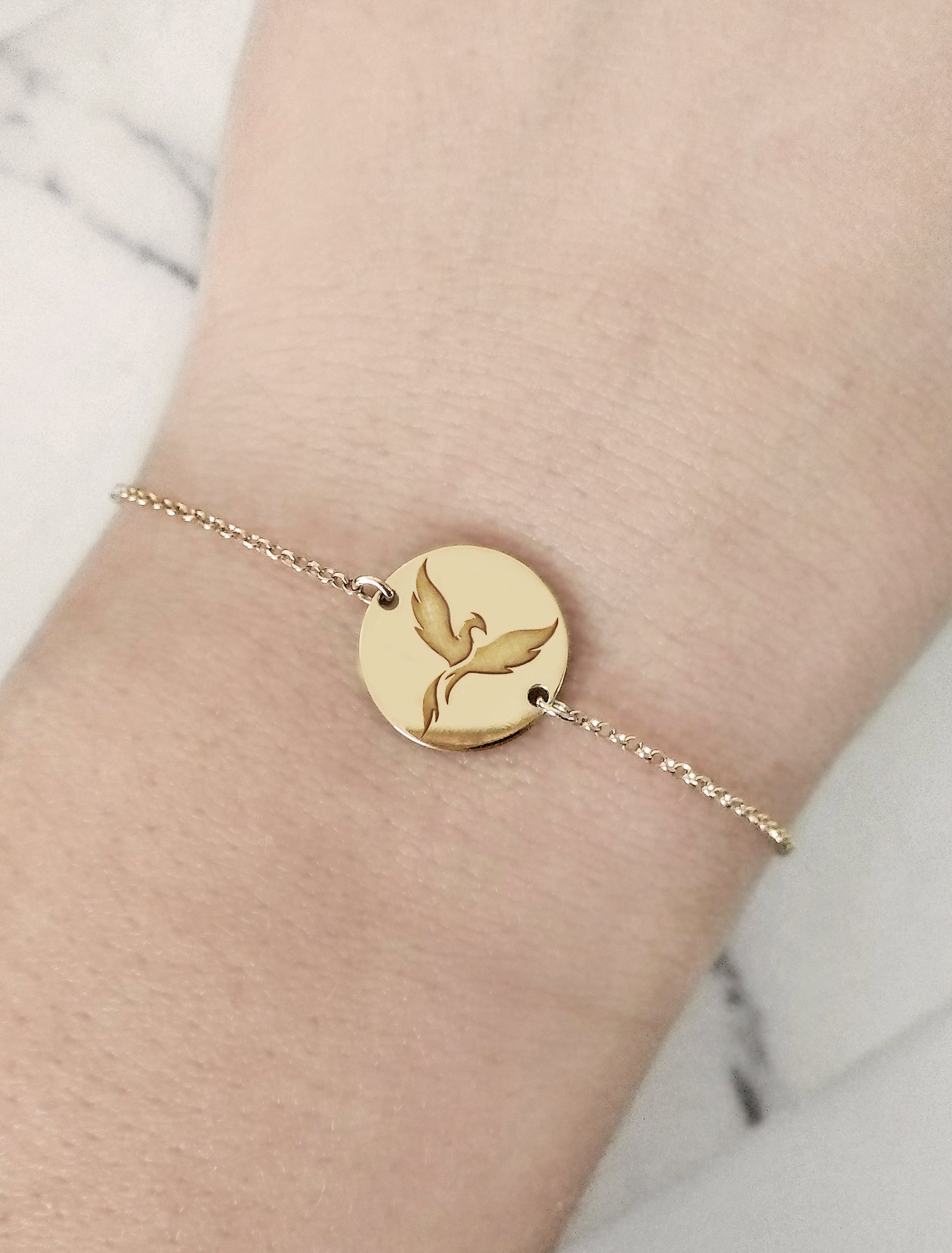 close up of woman's wrist wearing a  gold bracelet with a 14mm disc charm engraved with a phoenix firebird rising