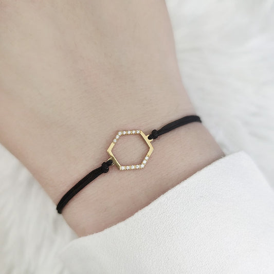Gold hexagonal charm bracelet with diamonds and black cord on a wrist against a white background