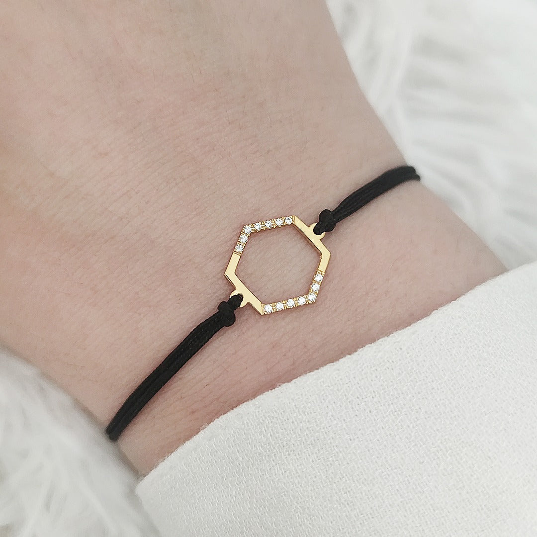 Gold hexagon charm bracelet set with diamonds with black cord on a wrist against a white background