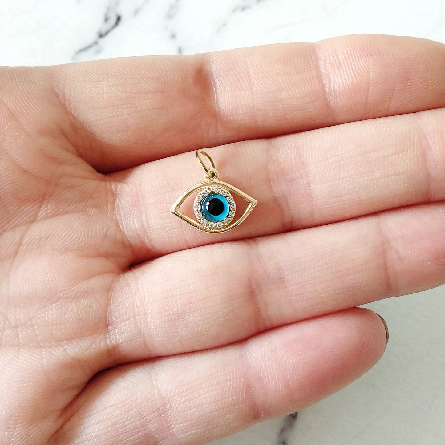 Gold eye-shaped pendant featuring a blue eye in the center surrounded by white diamonds held in a hand