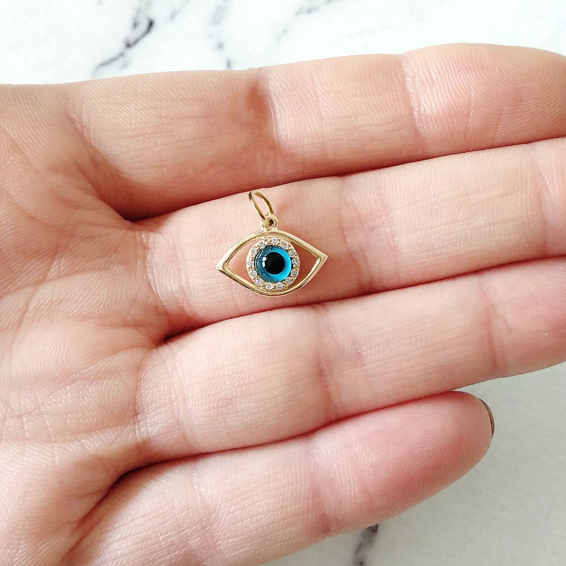 Gold eye-shaped pendant featuring a blue eye in the center surrounded by white diamonds held in a hand