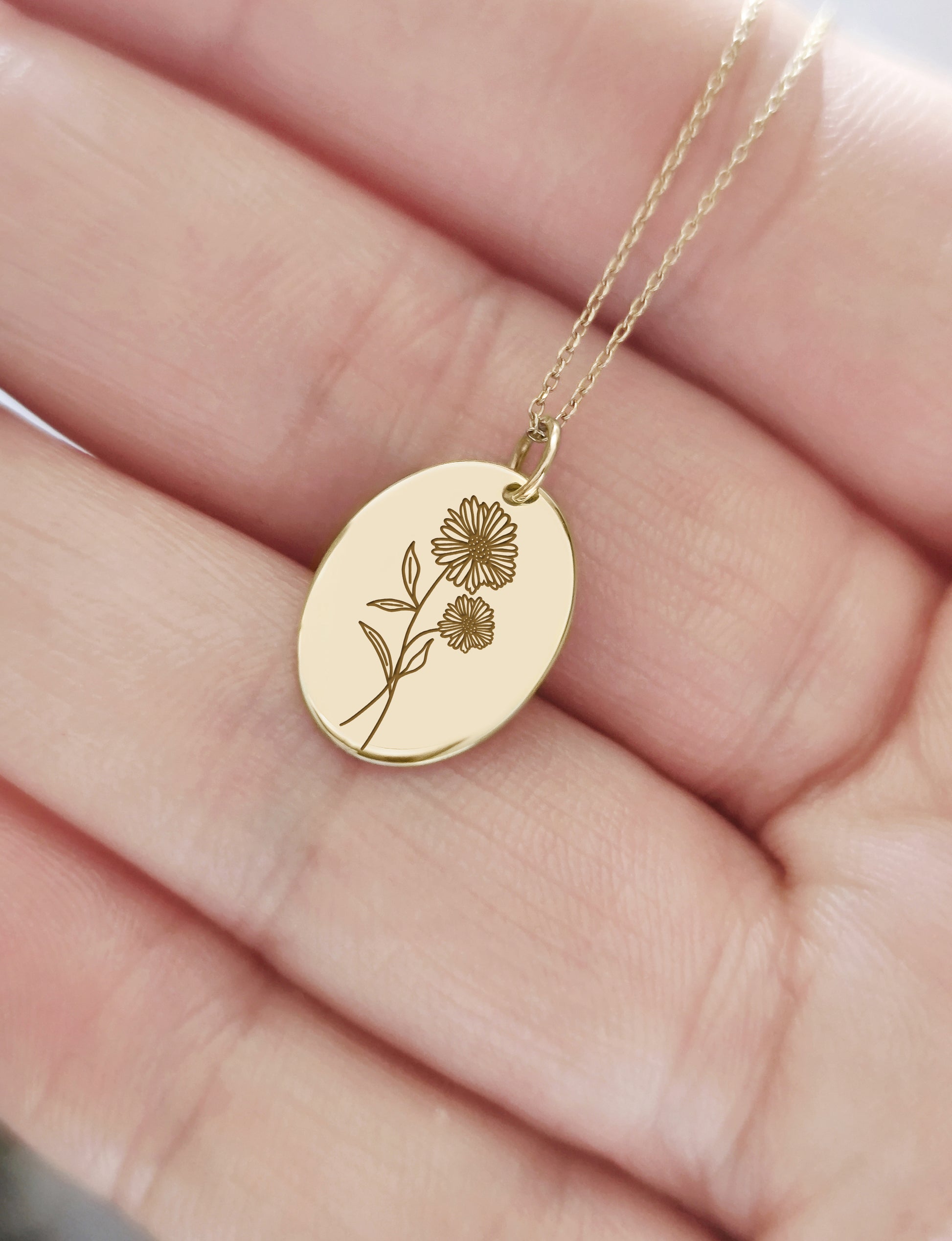 hand holding yellow gold oval pendant necklace engraved with the birth flower of April Daisy for size reference