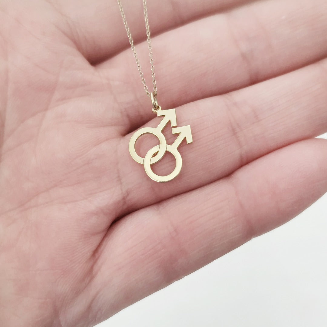 A gold necklace with a small double mars gay symbol pendant held on a palm