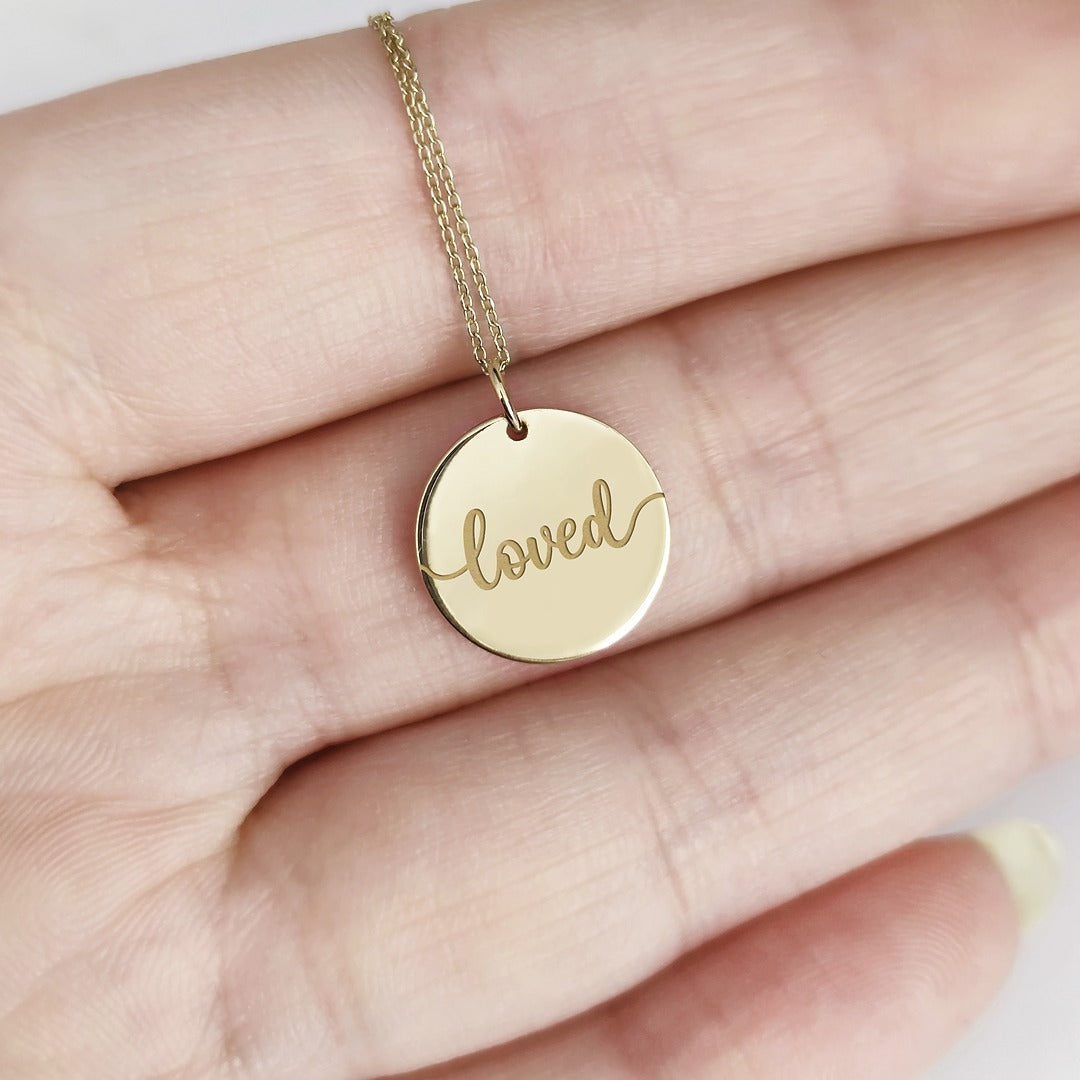 Gold necklace with a round pendant engraved with the word 'loved' held in a hand.