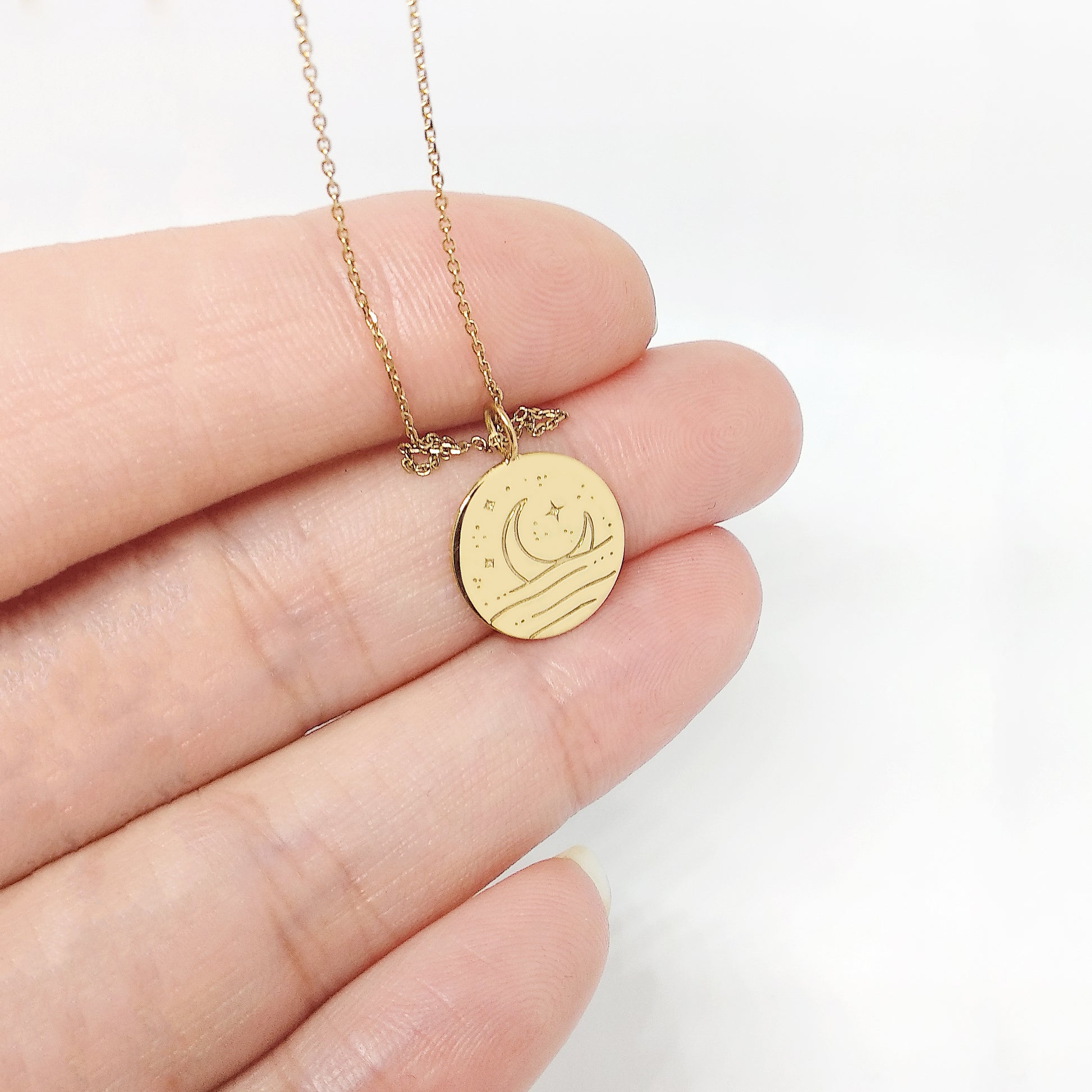 Gold necklace with a round pendant featuring a moon coming out of the sea held by a hand on a white background