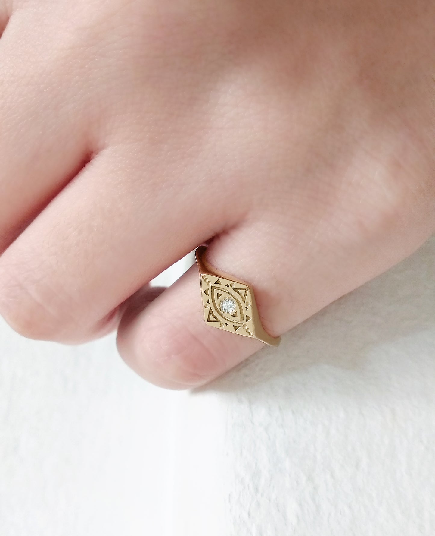 woman's pinky finger wearing gold rhombus signet ring engraved with a geometric evil eye and set with a tiny white diamond in the center
