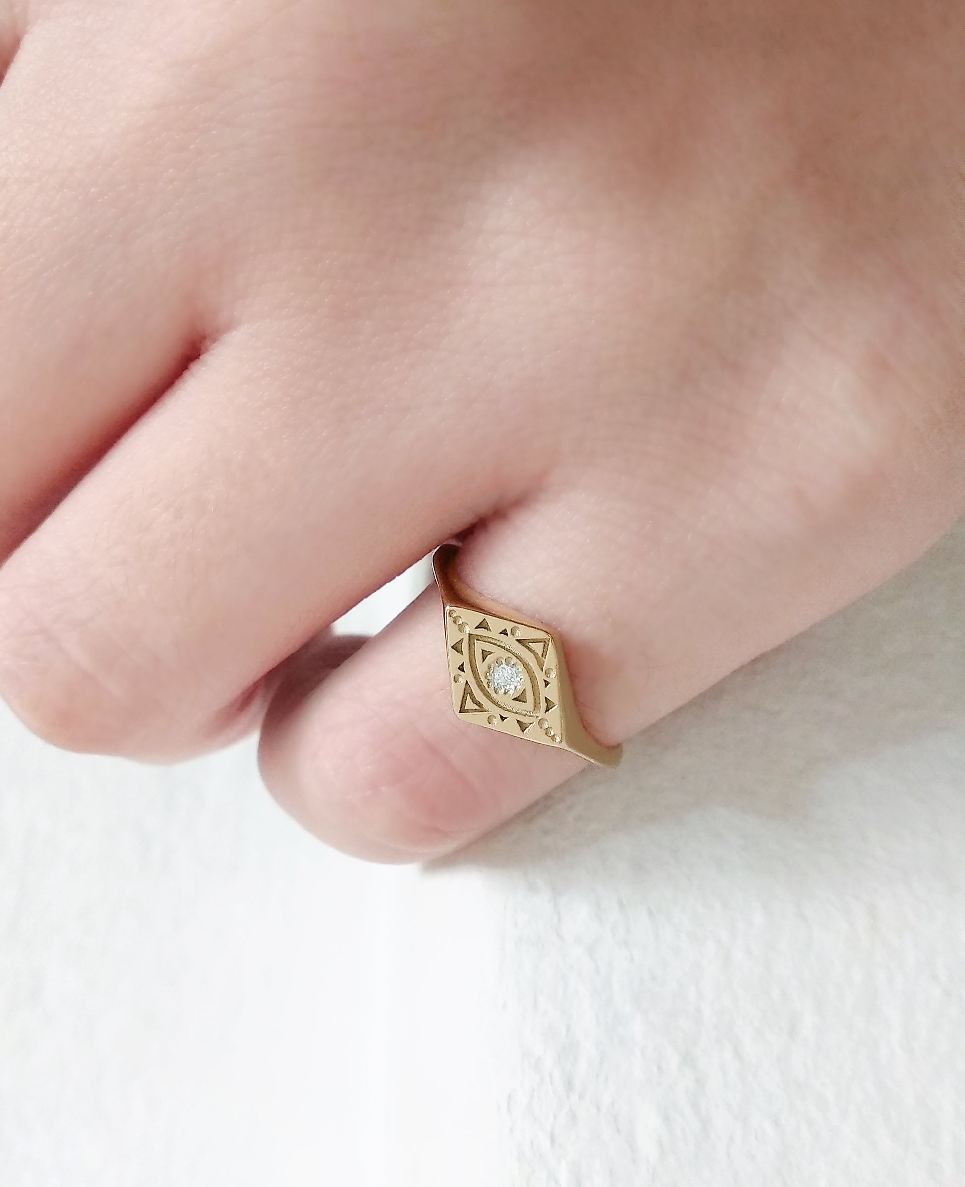 woman's pinky finger wearing gold rhombus signet ring engraved with a geometric evil eye and set with a tiny white diamond in the center