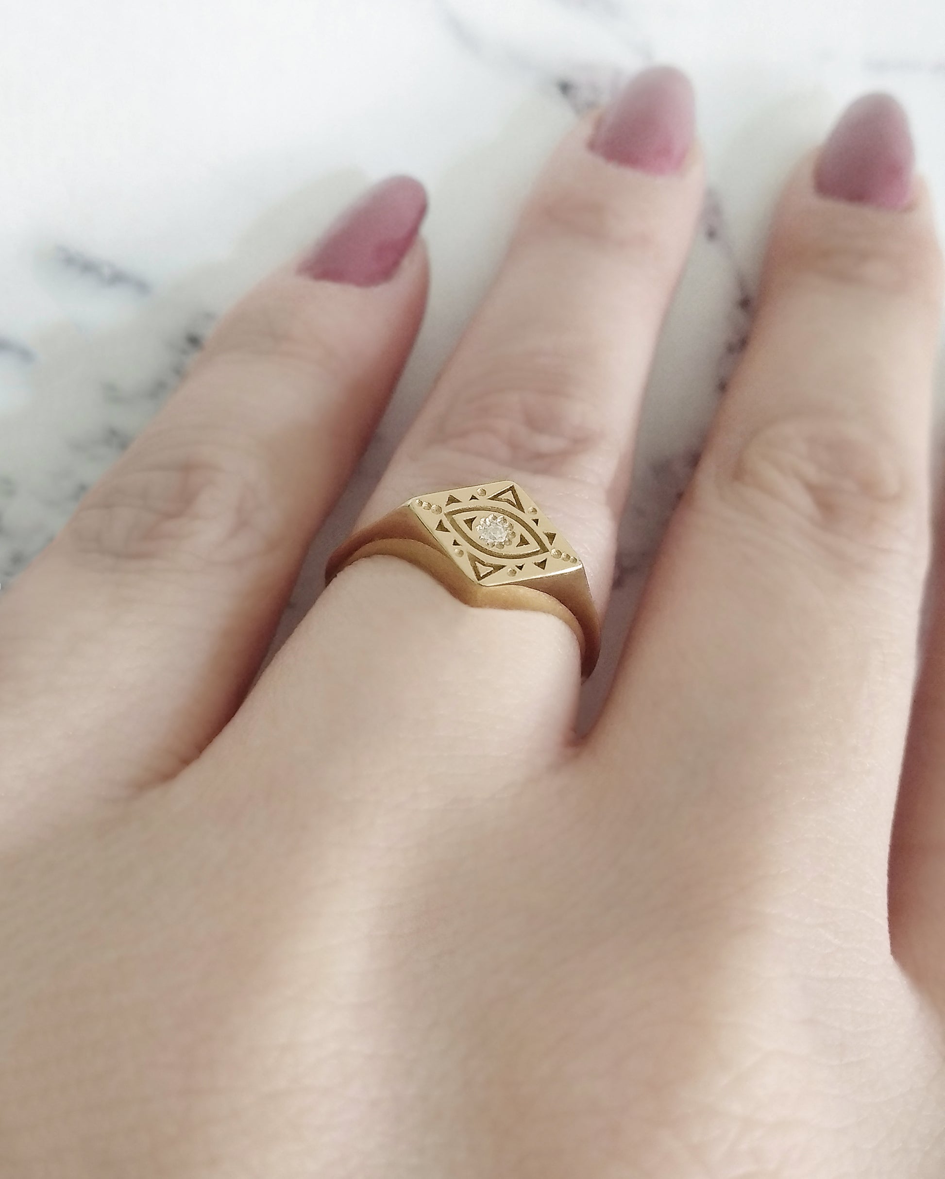 woman's ring finger wearing yellow gold rhombus signet ring engraved with a geometric evil eye and set with a tiny white diamond in the center