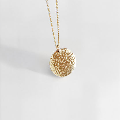 Gold necklace with a round, hammered pendant on a light gray background