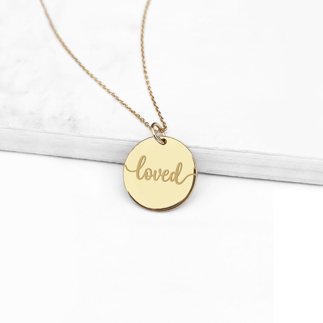 Gold necklace with a round pendant that says 'loved' on a white background